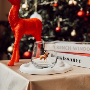 May include: A festive holiday scene featuring a red reindeer figurine and a clear glass with a miniature reindeer inside. The glass sits on a white, cloud-shaped base, with a Christmas tree in the background.