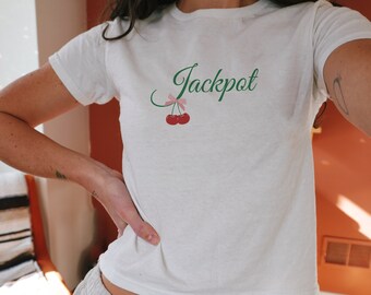 May include: White crop top with the word "Jackpot" in green script, a pink bow, and two red cherries. The top is paired with light gray sweatpants with a drawstring.