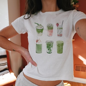 May include: White t-shirt with a graphic of six different green drinks with whipped cream, strawberries, and boba.