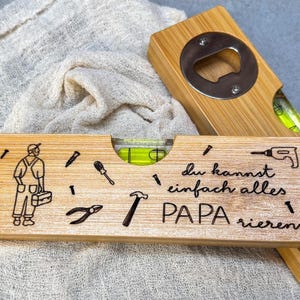 Könnte beinhalten: Holz-Wasserwaage und Flaschenöffner-Set. Die Wasserwaage zeigt eine Zimmermann-Illustration, Werkzeuge und den Text "du kannst einfach alles PAPA rieren". Der Flaschenöffner hat einen Metallöffner.
