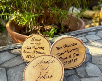 May include: Three wooden save the date signs with engraved text. The first sign says "Save our date Verena & Florian 02/07/" The second sign says "Wir Heiraten Tara & Eric 10/05/25". The third sign says "Amelie und Lukas 01.08.2024".