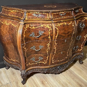 May include: A brown antique chest of drawers with a curved front and ornate details. The chest features multiple drawers with decorative handles and gold-colored accents. The legs are carved, and the overall design is elegant and classic.