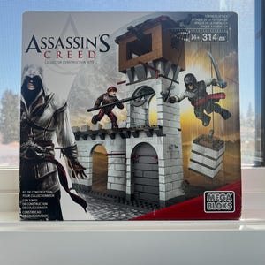 Op de afbeelding: Assassin's Creed Collector Construction Sets doos met een fortscène en figuren. De doos heeft het Assassin's Creed logo en de woorden "Fortress Attack". De set bevat 314 stukjes en is voor kinderen vanaf 14 jaar.