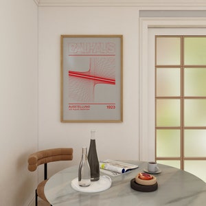 Pode incluir: Um pôster emoldurado com um design geométrico vermelho e branco. O pôster diz "BAUHAUS AUSSTELLUNG 16. August - September 1923". O pôster está pendurado em uma parede branca acima de uma mesa com um copo de água, uma garrafa de vinho e uma revista.