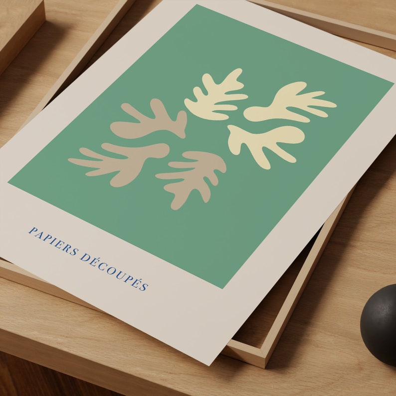 K&ouml;nnte beinhalten: Ein gerahmter Druck mit gr&uuml;nem Hintergrund und einem Design aus f&uuml;nf abstrakten Formen in Beige und Hellgelb. Der Text "PAPIERS D&Eacute;COUP&Eacute;S" ist unten auf dem Druck in Blau gedruckt.