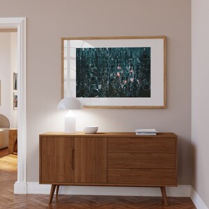 May include: A wooden sideboard with two doors and three drawers. The sideboard is a mid-century modern style with tapered legs. A white lamp and a bowl are on top of the sideboard. A framed abstract art print with a teal and pink color scheme hangs on the wall above the sideboard.
