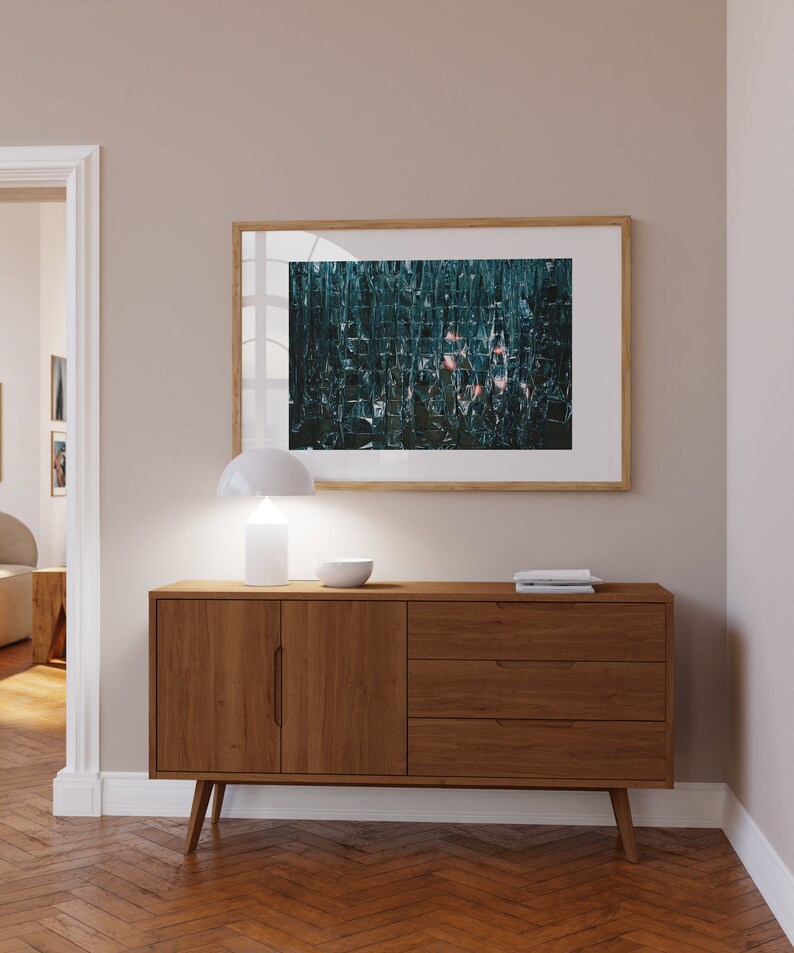 May include: A wooden sideboard with two doors and three drawers. The sideboard is a mid-century modern style with tapered legs. A white lamp and a bowl are on top of the sideboard. A framed abstract art print with a teal and pink color scheme hangs on the wall above the sideboard.