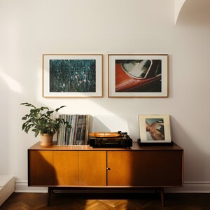 May include: A mid-century modern wooden cabinet with two doors and a record player on top. The cabinet is in a room with two framed photographs of abstract art and a photograph of a car. A potted plant sits on the cabinet.