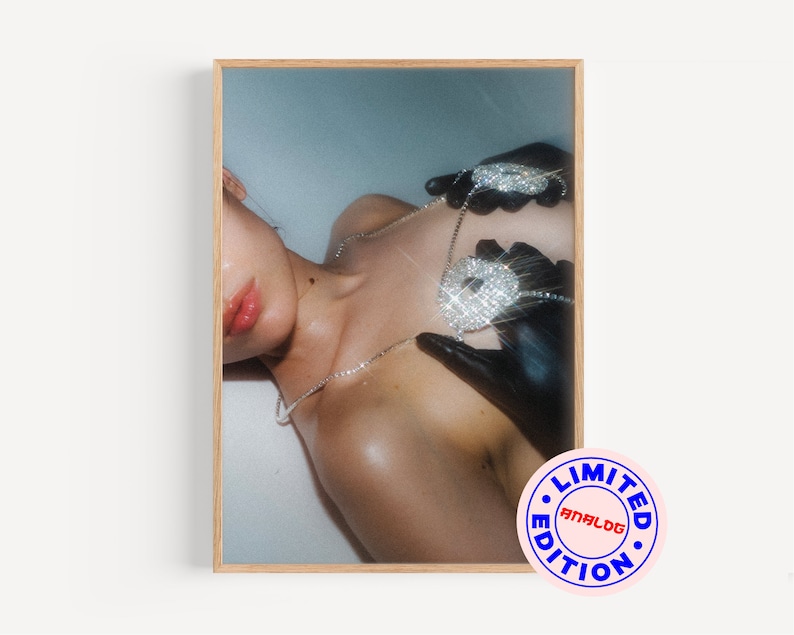 Op de afbeelding: Een close-up van de borst van een vrouw met een grote, met diamanten bezette ketting met hanger en zwarte handschoenen. De ketting heeft de vorm van een dollar-teken. De achtergrond is lichtblauw. De afbeelding heeft een "LIMITED EDITION ANALOG" stempel in de rechterbenedenhoek.