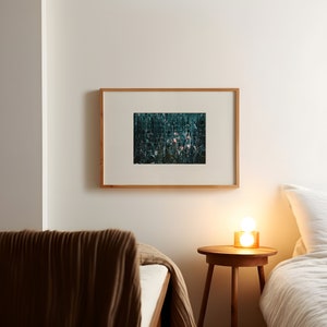 May include: A framed print of a dark green abstract design with a few pink accents hangs on a white wall above a nightstand with a lamp and a bed with a brown blanket.