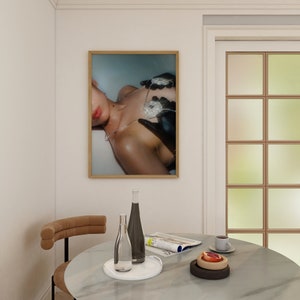 Op de afbeelding: Een ingelijste foto van een persoon die een met diamanten bezette ketting en handschoenen draagt. De foto hangt aan een witte muur boven een tafel met een glazen fles, een tijdschrift en een bord met fruit.