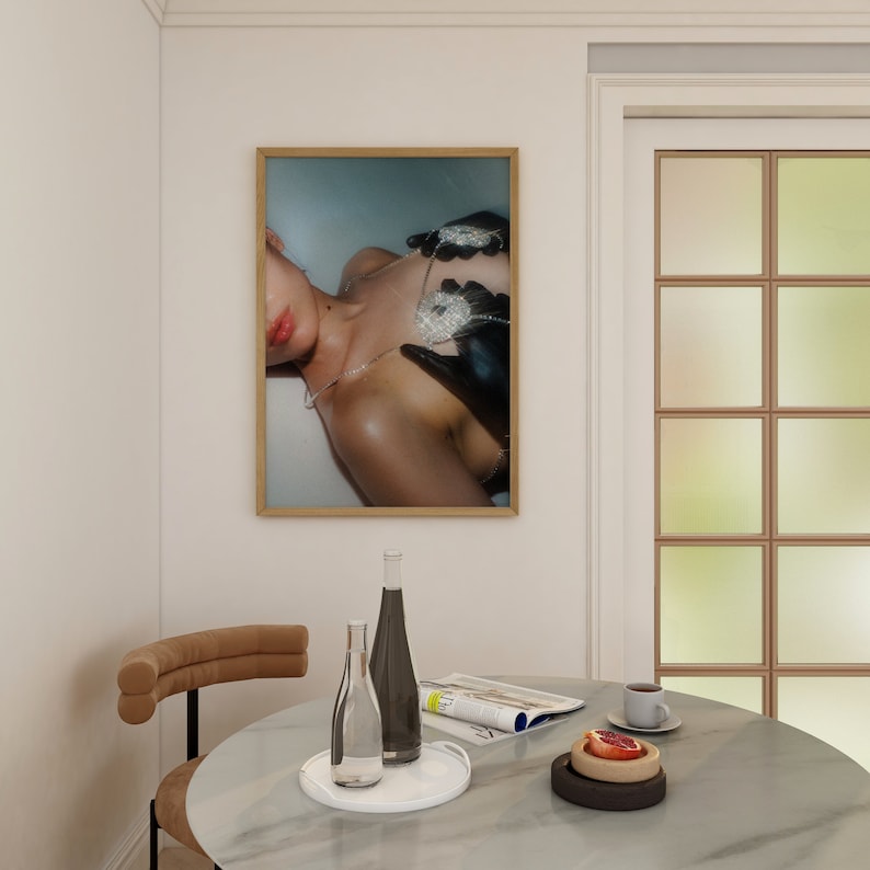 Op de afbeelding: Een ingelijste foto van een persoon die een met diamanten bezette ketting en handschoenen draagt. De foto hangt aan een witte muur boven een tafel met een glazen fles, een tijdschrift en een bord met fruit.