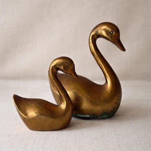 May include: Two vintage brass swan figurines. The larger swan has a long, curved neck and a detailed head. The smaller swan is in a similar pose. Both are gold-toned and sit on a neutral surface.