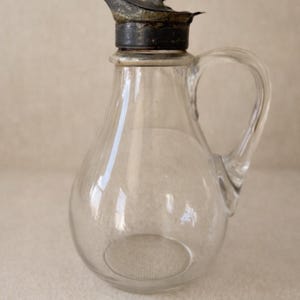 May include: A clear glass pitcher with a rounded body and a curved handle. The pitcher has a metal lid with a spout and a small decorative figure on top. The pitcher is likely used for serving liquids.