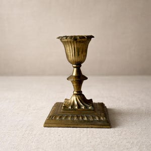 May include: A vintage brass candlestick holder with a square base and a fluted, tulip-shaped cup. The candlestick is approximately 6 inches tall and has an ornate, detailed design.