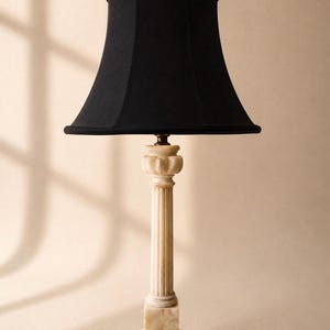 May include: A table lamp featuring a black fabric bell-shaped lampshade and a base crafted from light beige, marbled stone. The lamp base is designed with a fluted column structure, a square pedestal, and a decorative carved top section.