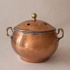 May include: A copper-colored pot with a lid and two handles. The lid has a small brass knob and three small holes. The pot has a rounded shape and is made of shiny copper. The handles are a brass color.