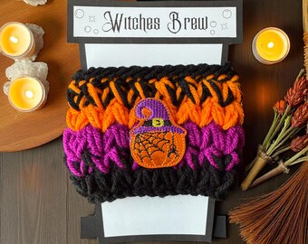 May include: A handmade, knitted coffee cup cozy in black, orange, and purple. The cozy features an embroidered patch of a witch's cauldron with a spider web and a witch's hat. The text "Witches Brew" is displayed on a sign above the cozy.