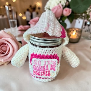 May include: A white and pink crocheted jar cozy with the text "SO IT'S GONNA BE FOREVER" and a ring graphic. The cozy is designed to fit around a jar, with a hat-like top and small arm-like extensions. The background includes pink roses.