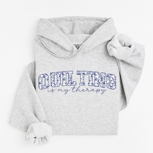 May include: A light gray hoodie with the text "QUILTING is my therapy" in blue floral lettering. The hoodie has a hood and long sleeves with ribbed cuffs. The design is centered on the front of the sweatshirt.