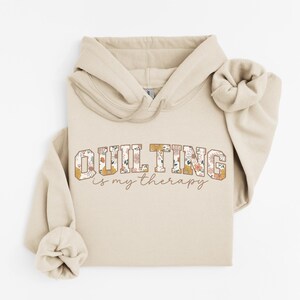 May include: A beige hoodie with the text "QUILTING is my therapy" in a floral design. The hoodie has a drawstring hood and long sleeves with rolled cuffs. The text is in a serif font.