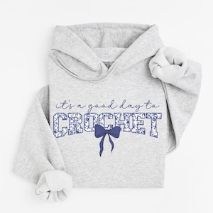 May include: A light gray hoodie with the phrase "it's a good day to CROCHET" in blue script and block letters. The word CROCHET is filled with a blue floral pattern, and a blue bow is below the word.