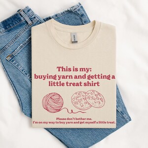 May include: A beige t-shirt with the text "This is my: buying yarn and getting a little treat shirt" in red. The text is accompanied by a drawing of a ball of yarn and two cookies.