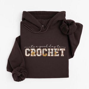 Puede incluir: Una sudadera con capucha marr&oacute;n con el texto "it's a good day to CROCHET" en blanco con acentos florales. La palabra "CROCHET" est&aacute; en letras grandes y en negrita.