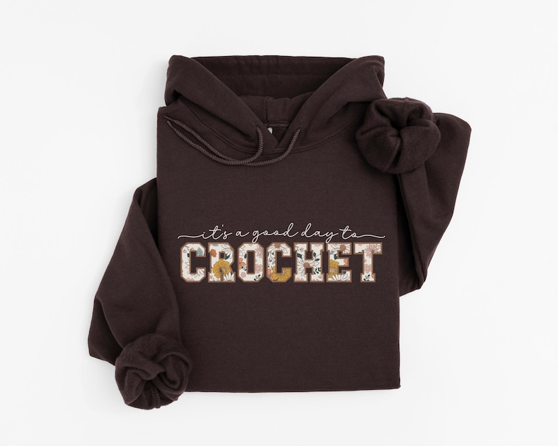 Puede incluir: Una sudadera con capucha marr&oacute;n con el texto "it's a good day to CROCHET" en blanco con acentos florales. La palabra "CROCHET" est&aacute; en letras grandes y en negrita.