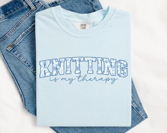 May include: A light blue t-shirt with the text "Knitting is my therapy" in a blue and white floral font.