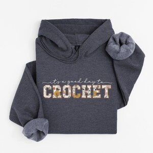 Puede incluir: Una sudadera con capucha gris oscuro con el texto "it's a good day to CROCHET" en blanco con acentos florales. El texto est&aacute; en una fuente de estilo vintage.