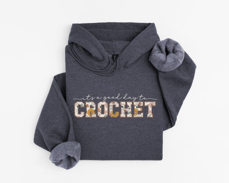Puede incluir: Una sudadera con capucha gris oscuro con el texto "it's a good day to CROCHET" en blanco con acentos florales. El texto est&aacute; en una fuente de estilo vintage.