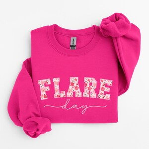 May include: A pink sweatshirt with the text "FLARE day" in white letters with a floral pattern. The letters are in a bold, blocky font.