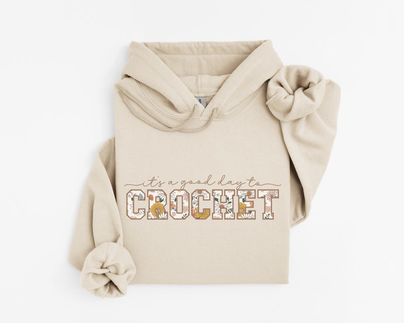Puede incluir: Una sudadera con capucha beige con un dise&ntilde;o floral que dice "es un buen d&iacute;a para tejer crochet" en texto marr&oacute;n.