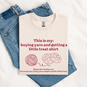 May include: A cream-colored t-shirt with the text "This is my: buying yarn and getting a little treat shirt" in red. Below the text are illustrations of a ball of yarn and two cookies.