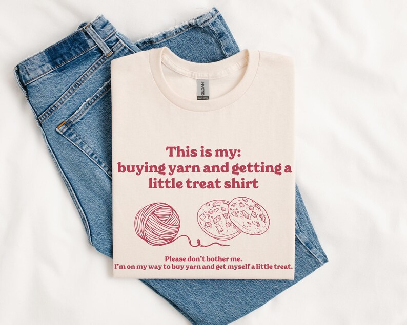 May include: A cream-colored t-shirt with the text "This is my: buying yarn and getting a little treat shirt" in red. Below the text are illustrations of a ball of yarn and two cookies.