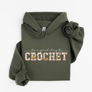 Puede incluir: Una sudadera con capucha verde con el texto "it's a good day to CROCHET" en blanco con detalles florales.
