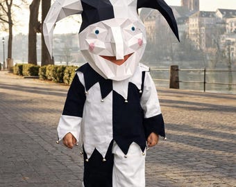Op de afbeelding: Een persoon in een zwart-wit harlekijnkostuum met een groot, geometrisch masker. Het kostuum omvat een bijpassende tuniek en broek, met belletjes aan de manchetten. De achtergrond toont een geplaveide loopbrug, bomen en gebouwen.