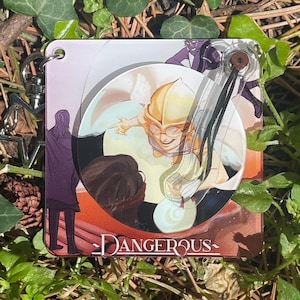 May include: A square acrylic keychain featuring a colorful illustration of a character with a golden helmet and glasses, with the word DANGEROUS printed at the bottom. The design includes a metallic star-shaped clasp and a small black cord attachment.