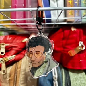 May include: A clear acrylic keychain featuring a stylized illustration of a person wearing orange-tinted glasses and a headset. The keychain is attached to a black metal clip, suitable for keys or bags. The illustration has a green and black color scheme.