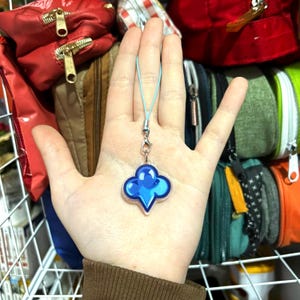 Pode incluir: Um pingente azul com um design de trevo de quatro folhas, preso a um cordão azul claro. O pingente é segurado em uma mão, com várias bolsas e bolsas no fundo.