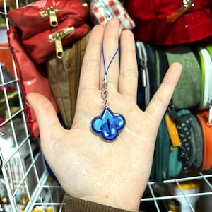 May include: A blue and teal charm with a four-leaf clover design, attached to a blue lanyard and silver clasp. The charm is held in front of a background of various bags and pouches.