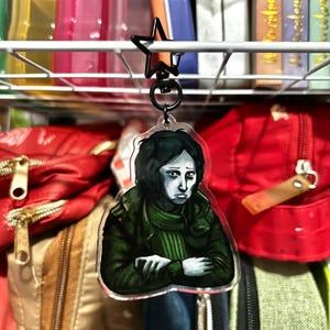 Op de afbeelding: Een heldere acryl sleutelhanger met een melancholische illustratie van een persoon in een groene jas en sjaal. De sleutelhanger heeft een zwarte stervormige clip. De achtergrond toont rode tassen en een wit draadrek.