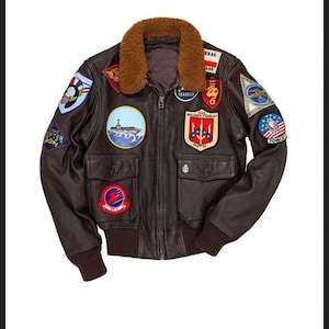 May include: A brown leather bomber jacket with a shearling collar and multiple patches. The patches include a variety of designs, such as a ship, an eagle, and a trident. The jacket has two pockets on the front and a zipper closure.