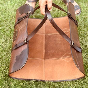 May include: A brown leather apron with adjustable straps. The apron is folded in half and has a dark brown leather trim.