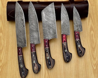 Puede incluir: Conjunto de cinco cuchillos de cocina con hojas de acero Damasco y mangos de madera. Los cuchillos están dispuestos en una fila sobre una superficie de madera. Los cuchillos tienen acentos rojos y negros en los mangos.