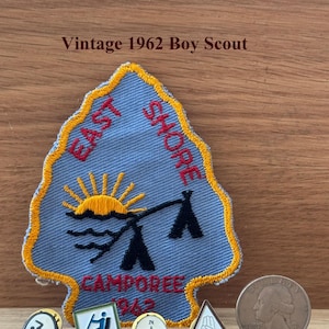 May include: Vintage 1962 Boy Scout memorabilia, including a blue and yellow patch with a sun and tent design, and several pins. A quarter is also present. The patch reads "East Shore Camporee 1962".