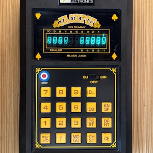 Könnte beinhalten: Schwarzes elektronisches Handheld-Spiel mit gelbem und schwarzem Design. Das Spiel verfügt über ein digitales Display mit der Aufschrift "JACKPOT GIN RUMMY" und eine Tastatur mit nummerierten Tasten. Der Markenname "INTEX ELECTRONICS" ist sichtbar.