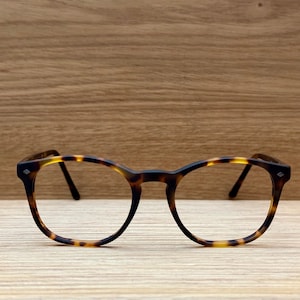 Puede incluir: Gafas de montura redonda con un patrón de carey. Las gafas tienen un puente y patillas de color marrón oscuro, con un acento en forma de diamante en las esquinas exteriores. Las lentes son transparentes y la montura está sobre un fondo de madera.