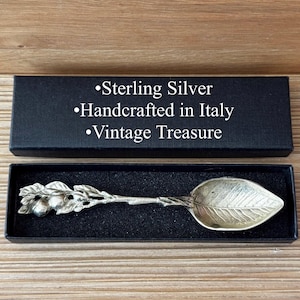 Puede incluir: Una cuchara de plata de ley con un cuenco en forma de hoja y un mango adornado con una rama y bayas, presentada en una caja negra. La caja dice "Sterling Silver Handcrafted in Italy Vintage Treasure."